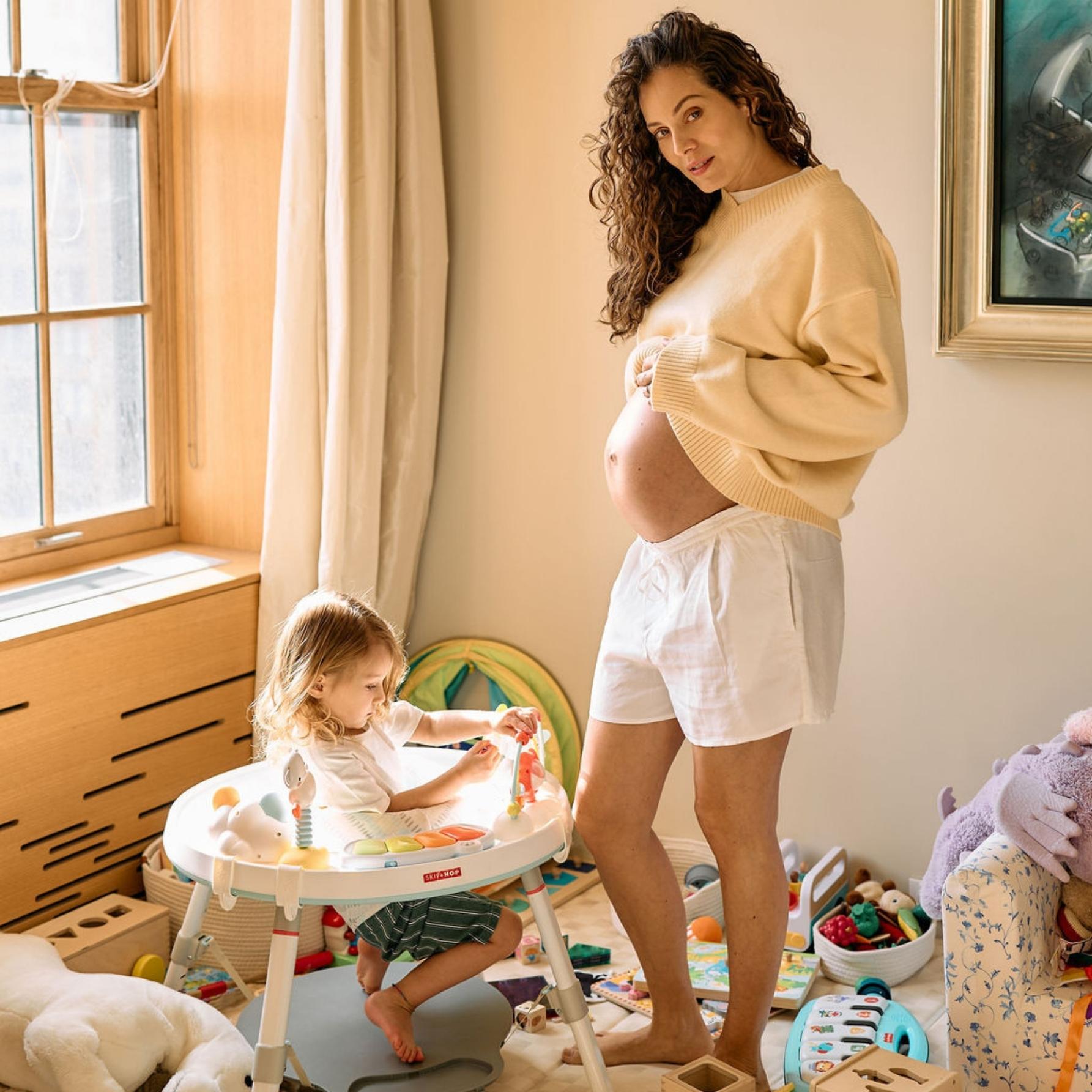 A pregnant woman stands barefoot in a playroom while holding her baby bump, next to a toddler playing. 